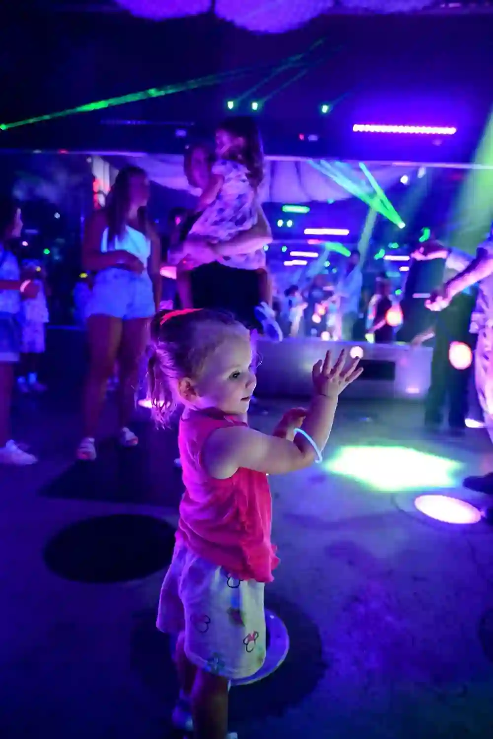 Toddler clapping on the glow-lit dance floor with families behind her